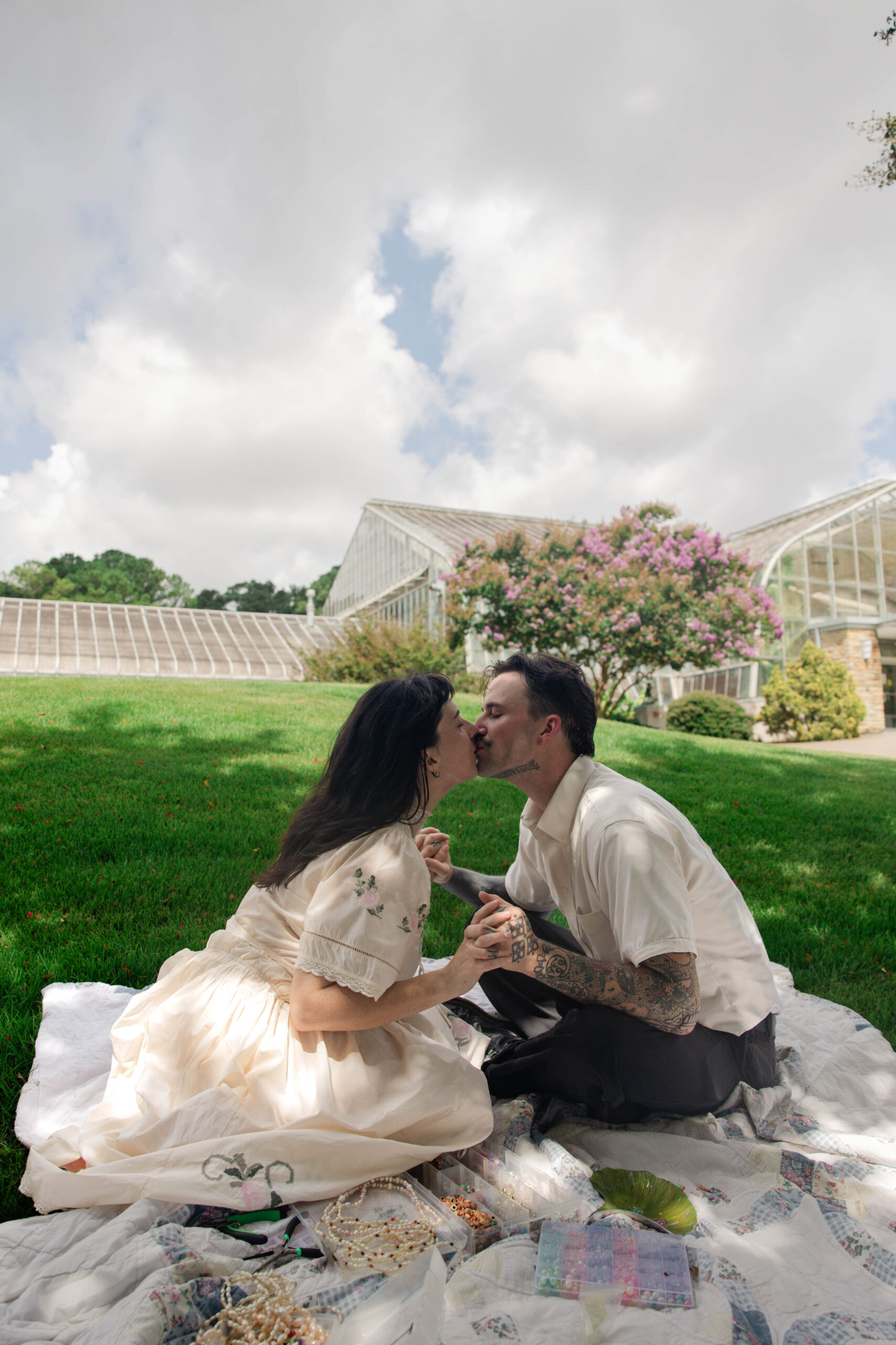 couple share a kiss on a picnic blanket with a green house standing behind them