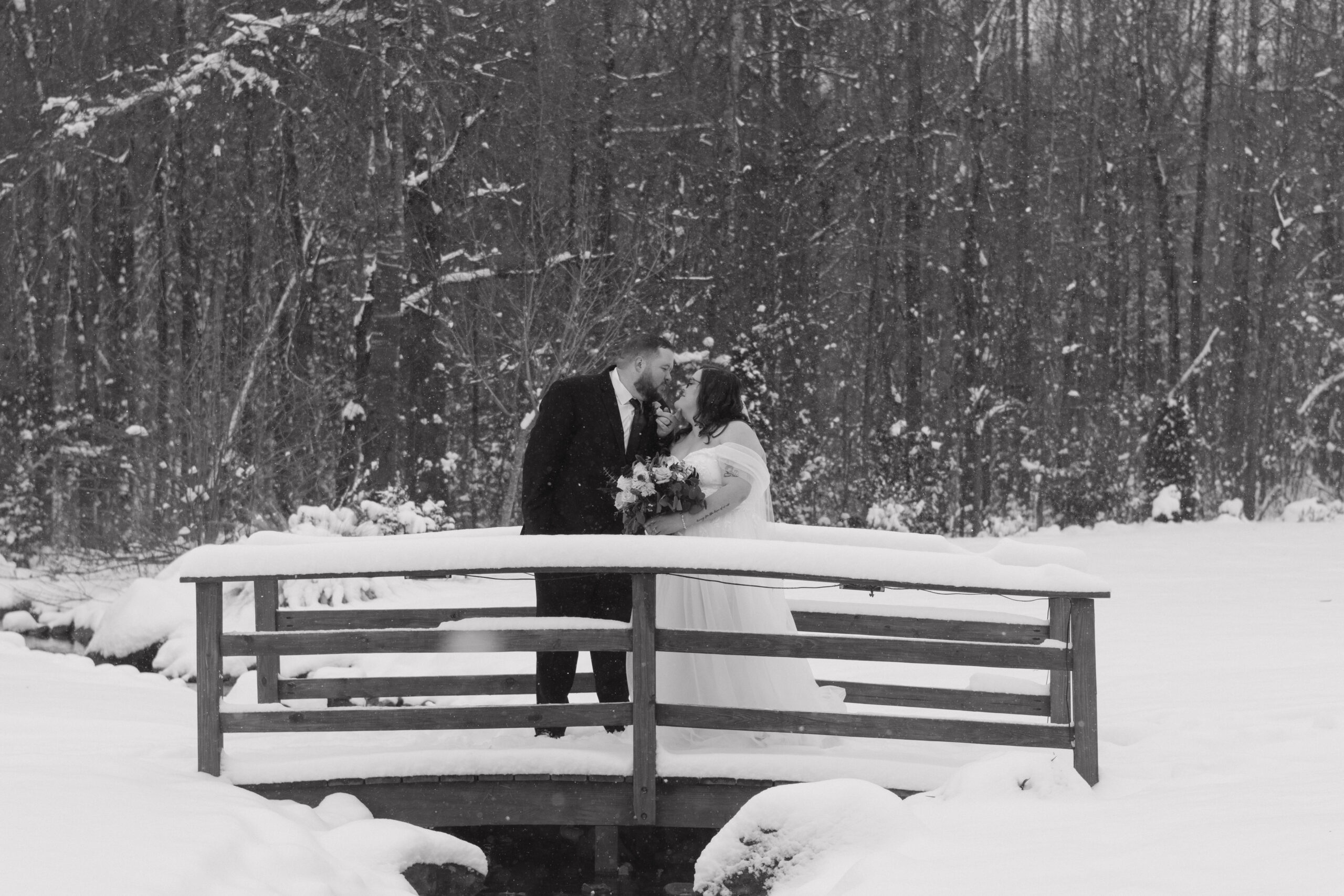 stunning couple share a kiss on a snow covered bridge during their white fox cottage wedding